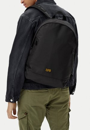 Person mit schwarzem Rucksack mit gelbem G-Star-Logo, schwarzer Jeansjacke und olivgrüner Cargohose, mit dem Rücken zur Kamera.