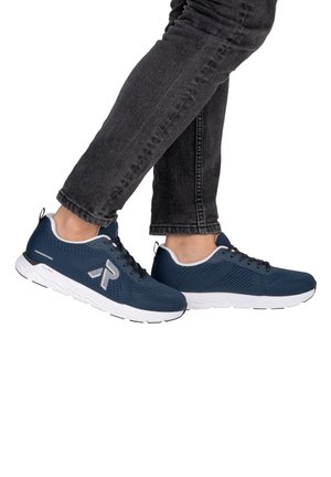 Osoba nosící tmavě modré tenisky s bílými podrážkami a černé džíny, nohy překřížené na bílém pozadí.