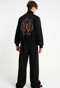 Czarna sportowa kurtka z wysokim kołnierzem, z kolorowym nadrukiem przedstawiającym dwa tygrysy oraz napisem "MMA" na plecach. Pasujące czarne spodnie.