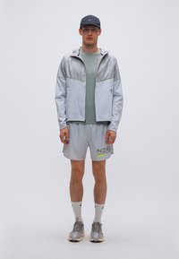 Αθλητικό μπουφάν ανοιχτού γκρι με γυαλιστερό ασημί πάνω μέρος, συνδυασμένο με πράσινο μπλουζάκι και γκρι σορτς που ταιριάζουν, διακοσμημένα με λογότυπο της Nike.