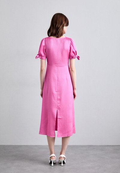 Rosa midiklänning med texturerad tyg, puffärmar knutna vid armbågarna och en slits bak. Stylad med vita klacksandaler.