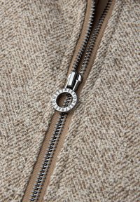 Brauner Stoff mit Struktur und einem Reißverschluss. Silberne Beschläge zeigen einen runden Anhänger mit der Aufschrift "STREET ONE". Eine Nahaufnahme hebt die Nähte und das Muster hervor.