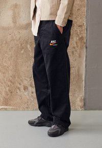 Black corduroy pants featuring a prominent orange Nike logo and text on the left leg, paired with dark athletic shoes.