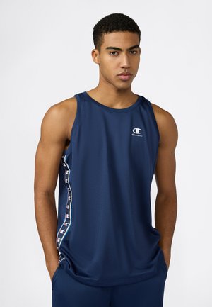 Giovane con capelli ricci corti che indossa una canotta sportiva blu navy senza maniche Champion e pantaloncini abbinati, in piedi con le mani in tasca.