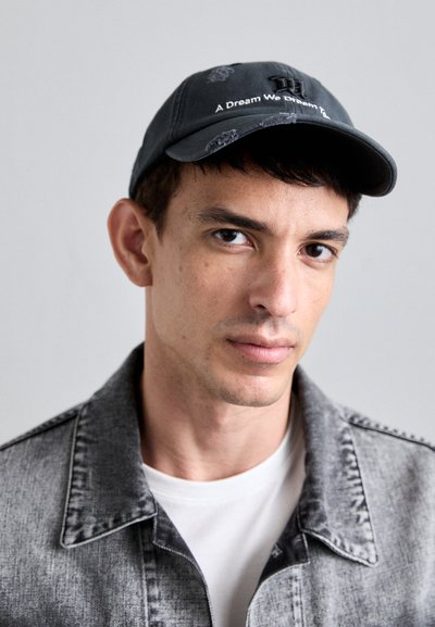 Black cap with curved brim, embroidered white text "A Dream We Dream To" on front, worn with a gray denim jacket and white shirt.