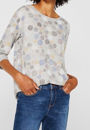 Kvinde iført lysegrå langærmet skjorte med flerfarvede mønstrede cirkler, kombineret med blå jeans, hænderne i lommerne, mod en ensfarvet baggrund.