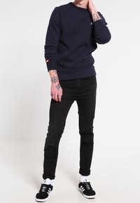 Persona che indossa una felpa blu navy, jeans skinny neri strappati, calze bianche Adidas e sneaker nere Adidas, in piedi su uno sfondo bianco.