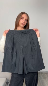 Sivé pruhované nohavice s vysokým pásom a záhybmi, s pútkami na opasok a bočnými vreckami, držiace model na neutrálnom pozadí.