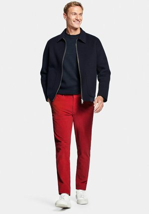 Man loopt, draagt een zwarte jas met rits, donkere trui, rode broek en witte sneakers op een effen lichte achtergrond.