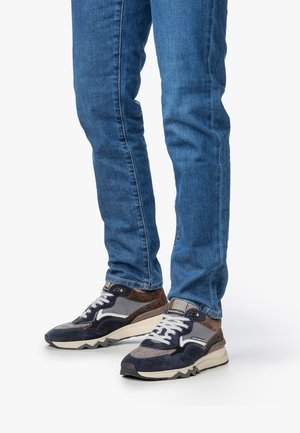 Jeans blu slim-fit indossati con sneakers lacci navy, grigie e marroni con suole bianche su uno sfondo bianco.
