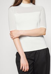 Femme portant un haut blanc côtelé à manches courtes avec des rayures horizontales transparentes et un pantalon noir, les bras croisés à la taille.