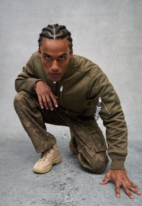 Chaqueta bomber verde oliva con bolsillos con cremallera, pantalones cargo de camuflaje y zapatillas beige. Tejido suave, corte relajado y detalles sutiles de logo.