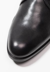 Chaussure en cuir noir avec une finition brillante, à bout pointu et au design épuré. Comprend un bord lisse et des détails matériels minimaux.