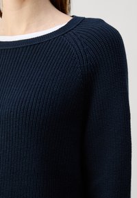 Sweater de malha azul-marinho com textura canelada, gola ampla em forma de barco e mangas raglan. Detalhe da área do ombro.