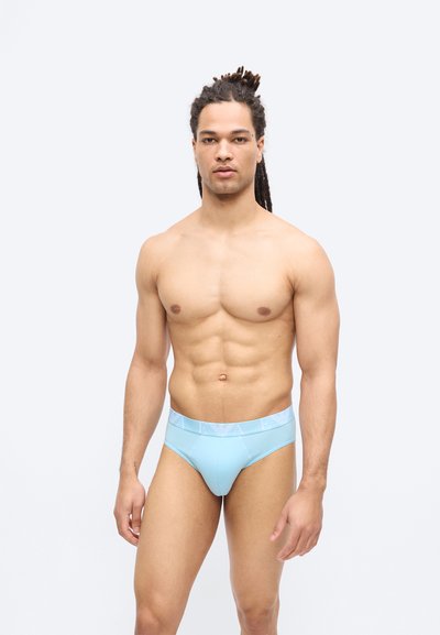 Κατάλληλος άνδρας μεν dreadlocks στέκεται με το πρόσωπο προς τα εμπρός φορώντας ανοιχτό μπλε εσώρουχα σε απλό φωτεινό φόντο.