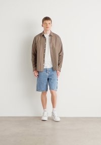 Hnědá košile s knoflíky přes bílém tričku, doplněná o světle modré denimové šortky a bílé tenisky, stojící na neutrálním pozadí.