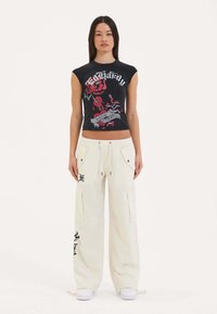 Zwarte mouwloze crop top met een grafisch ontwerp van een rode roos en tekst; gecombineerd met crèmekleurige baggy cargo broeken met zijzakken en zwarte accenten.