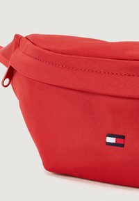Red fabric fanny pack with a smooth texture, zip closure, and a small embroidered logo featuring blue, white, and red stripes.