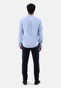 Camicia azzurra a maniche lunghe con orlo curvato e polsini abbottonati, abbinata a pantaloni scuri e scarpe nere, vista da dietro.