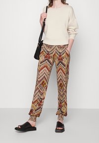 Beżowa bluza z długimi rękawami zestawiona z wielokolorowymi, wzorzystymi spodniami z falistymi i diamentowymi wzorami. Czarne klapki.
