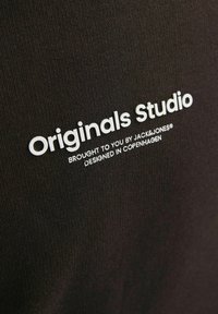 Černá látka s bílým textem, který zní "Originals Studio" a "Zajišťuje JACK&JONES" v čistém, moderním písmu.