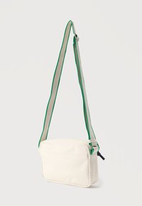 Λευκή τσάντα ώμου από καμβά με πράσινο και μπεζ λουράκι, ορθογώνιο σχήμα, φερμουάρ στο πάνω μέρος και μίνιμαλ σχεδιασμό. Πράσινες λεπτομέρειες.