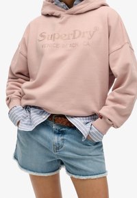 Sudadera rosa con el texto en relieve "SuperDry Venice Beach, CA", superpuesta a una camiseta de rayas azul y blanca, combinada con pantalones cortos de mezclilla desgastados.