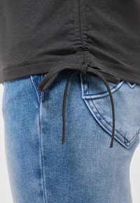 Čierna košeľa so zaväzovacím šnúrkou na spodnom okraji, nosená cez svetlomodré džínsové rifle s vreckom.