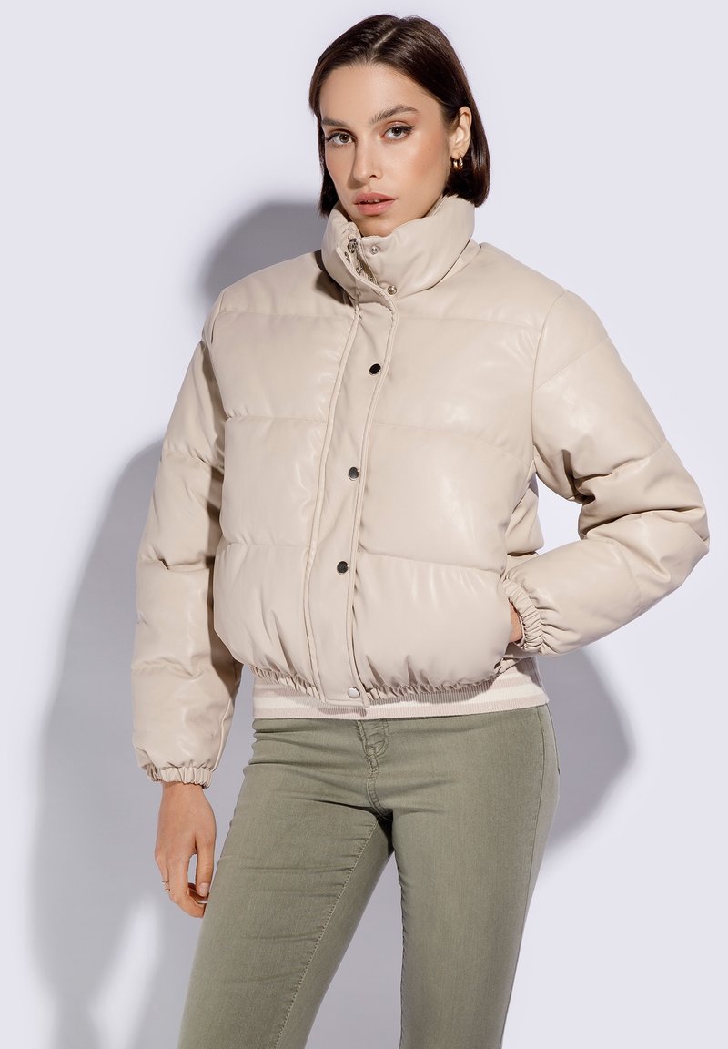 Beige puffervest med høj krave, trykknapper, elastiske manchetter og en tætsiddende talje. Glat, skinnende kunstlædertekstur.