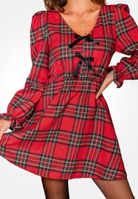 Rode geruite jurk met pofmouwen, V-hals en zwarte lintaccenten. Heeft een geraffineerde taille en een uitlopende rok. Zachte stoftextuur.