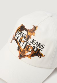 Bijela bejzbol kapa s istaknutim grafičkim dizajnom zlatnih i narančastih ukrasnih motiva te natpisom "VERSACE JEANS COUTURE" u podebljanoj crnoj boji.