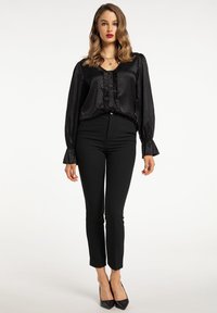 Sort struktureret bluse med rynker og knapper, parret med tætsiddende sorte bukser og spidse sorte hæle. Modellen står foran en neutral baggrund.