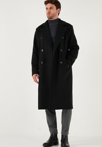 Melns vilnas maisījuma dubultā slēgšanas mētelis ar izgrieztiem atlokiem, sānu kabatām un teksturētu apdari, uzvilkts uz tumša turtleneck un pelēkiem biksēm.