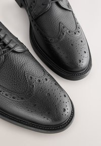 Chaussures habillées en cuir noir à finition texturée, design à bouts ailés et perforations décoratives sur le bout et les côtés.