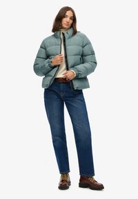 Polstret teal jakke med høj krave, frontlynlås og elastiske manchetter. Parret med blå jeans og brune snørebåndsstøvler.