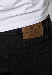 Blugi din denim negru cu o etichetă din piele ce prezintă logo-ul "MCS" și text. Detalii vizibile ale cusăturii și o nuanță de roșu pe marginea buzunarului.