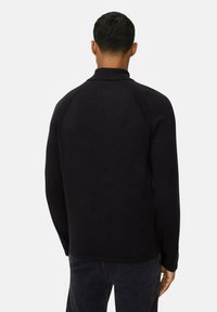 Mann von hinten gesehen, der einen schwarzen Langarmpullover und dunkle Hosen vor einem schlichten hellen Hintergrund trägt.