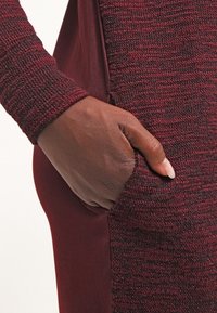 Abito bordeaux con maniche a maglia lavorata e tasca laterale, realizzato in tessuto liscio lungo il corpo e con una silhouette aderente.