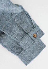 Helles blaues Denim-Hemd mit langen Ärmeln, Manschette mit einem einzelnen braunen Knopf und strukturiertem Stoff, der ein kontrastierendes Stickmuster aufweist.