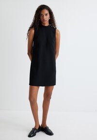Schwarzes ärmelloses Kleid mit einem hohen Kragen, geradem Schnitt und knielanger Länge. Glatter Stoff mit subtiler Textur. Tragen mit schwarzen Slippern.