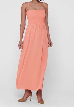 Woman wearing a sleeveless, ankle-length coral dress with smocked bodice and nude high-heeled shoes, standing against a plain background.