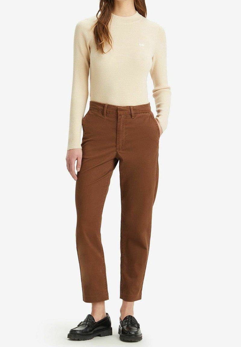 Beżowa bluzka z długim rękawem w prążki, zestawiona z brązowymi spodniami z bocznymi kieszeniami. Całość dopełniają czarne mokasyny. Prosty, estetyczny design.