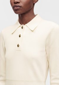 Personne portant un haut en maille crème avec col et trois boutons dorés sur le devant, présenté du cou à la taille.