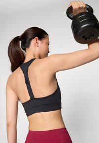 Soutien-gorge de sport noir avec des bretelles ajustables, présentant une texture lisse et un design dos nageur, associé à des leggings d'entraînement rouges.