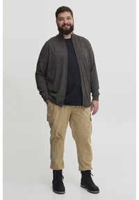Cárdigan gris oscuro con cierre frontal, usado sobre una camiseta negra, combinado con pantalones cargo caqui y botas negras con cordones. Diseño de atuendo casual.