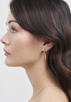 Young woman with brown wavy hair wearing a gold hoop earring with a silver dagger charm and a thin gold chain necklace.