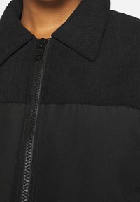 Gros plan sur une veste noire avec un col côtelé et une fermeture éclair visible à l'avant, portée par une personne à la peau de ton moyen.