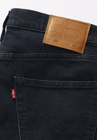 Dark denim jeans with a leather brand patch displaying "Levi Strauss & Co." and a red tag on the side. Features back pockets and stitching details.