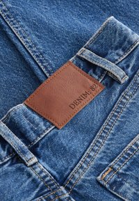 Traperice s plavom tkaninom i svijetlim pranjem. Smeđi kožni oparak s natpisom "DENIM: 81" ušiven na stražnjem dijelu struka.