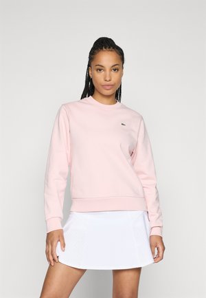 Woman with braided hair wearing a light pink sweatshirt and white perforated tennis skirt, standing against a plain gray background.